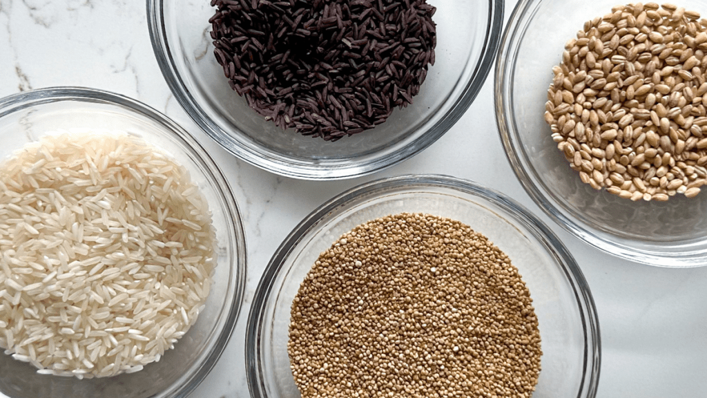 Four glass bowls containing raw ingredients for a healthy rice blend: white long-grain rice, quinoa, purple riceberry, and barley grains.