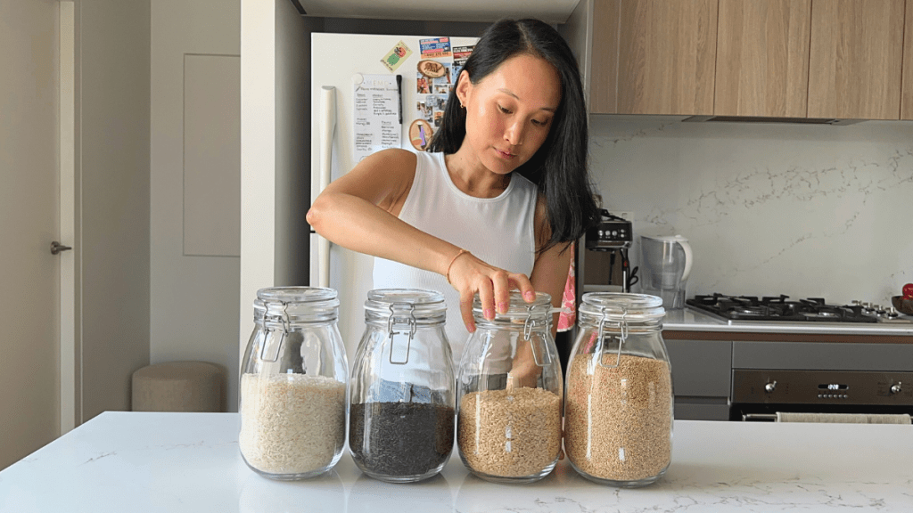 Woman arranging four large glass jars containing white rice, purple riceberry, brown rice, and quinoa on a white kitchen island.