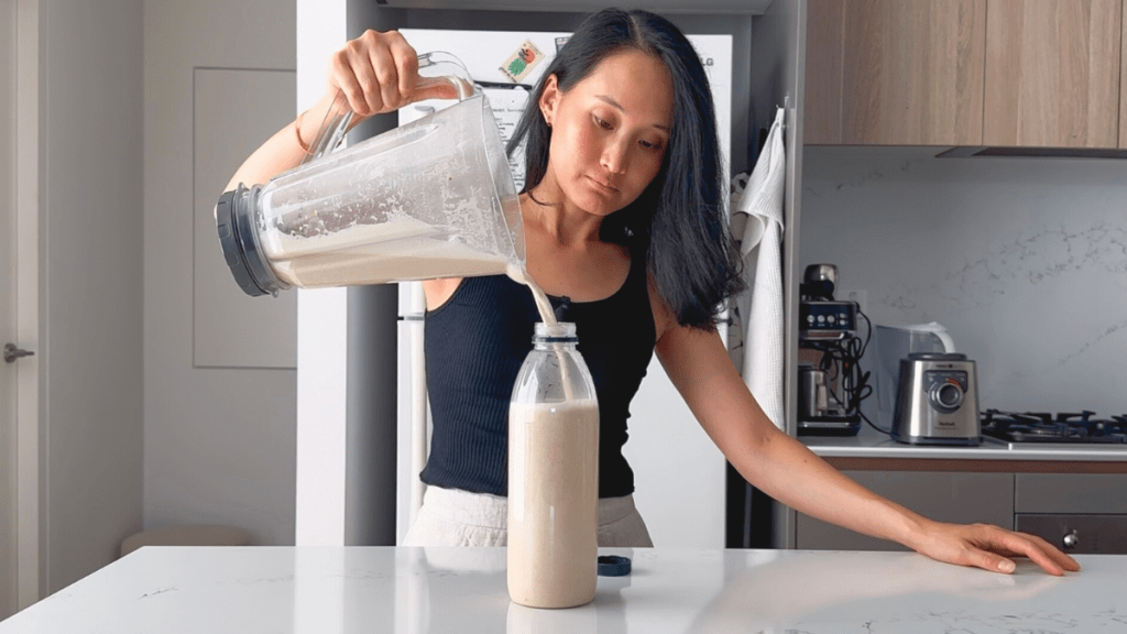 A person pouring creamy white cashew milk from a blender into a clear bottle.