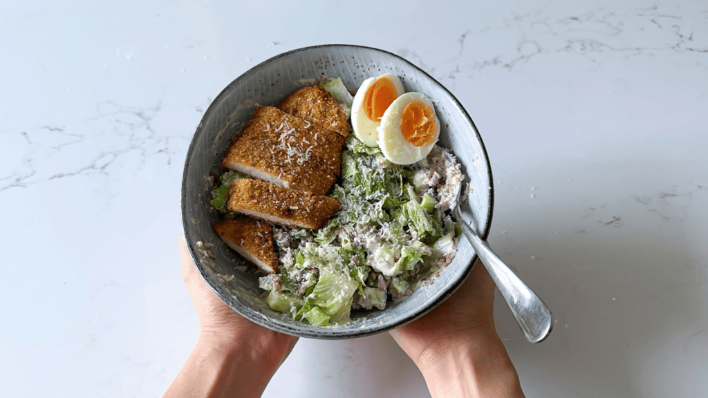 A healthy bowl featuring sliced air fryer chicken schnitzel, a boiled egg, chopped lettuce and cucumber, rice blend, and creamy garlic yoghurt dressing.