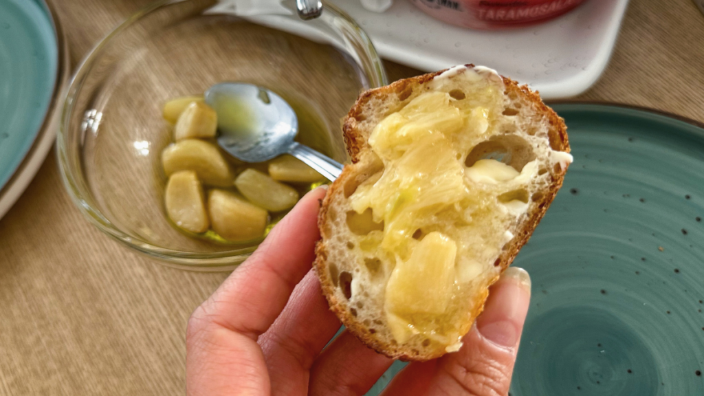 A close up shot of a person holding a slice of crusty bread topped with creamy, mashed roasted garlic confit.