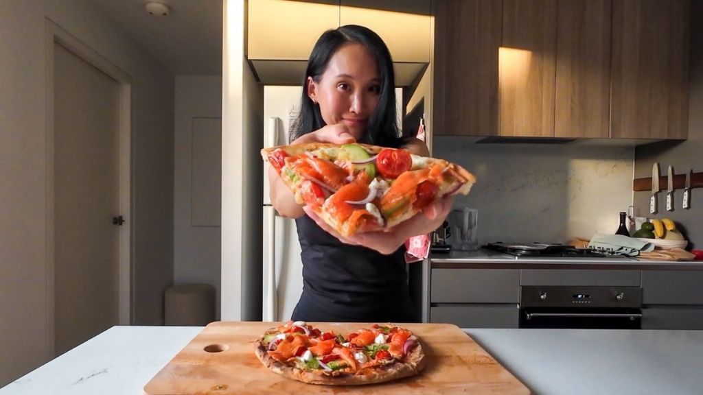 A woman in a kitchen holding a large, fresh slice of smoked salmon pizza topped with avocado and cherry tomatoes toward the camera.