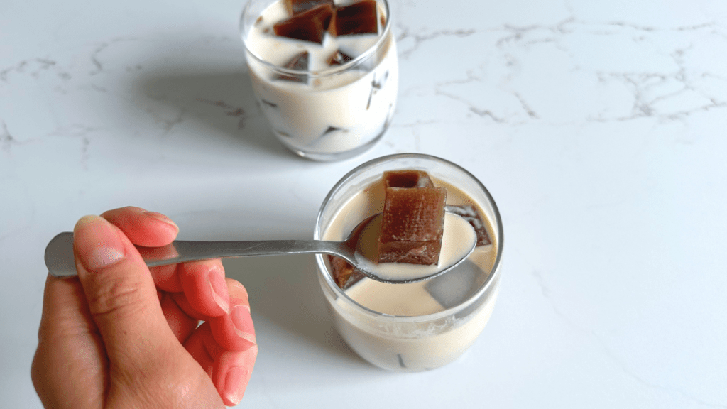 A close-up of a silver spoon lifting a single dark, translucent coffee jelly cube out of a glass of milk.