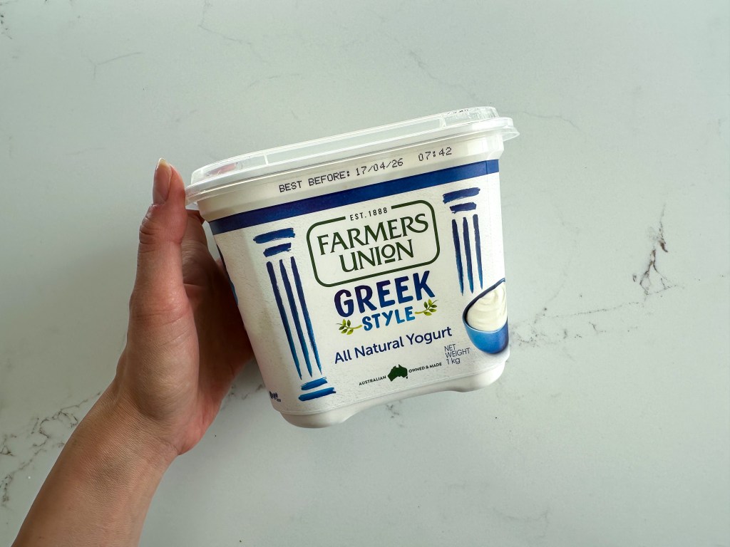 A hand holding a tub of Farmers Union Greek Style All Natural Yogurt to be used as a pizza base.