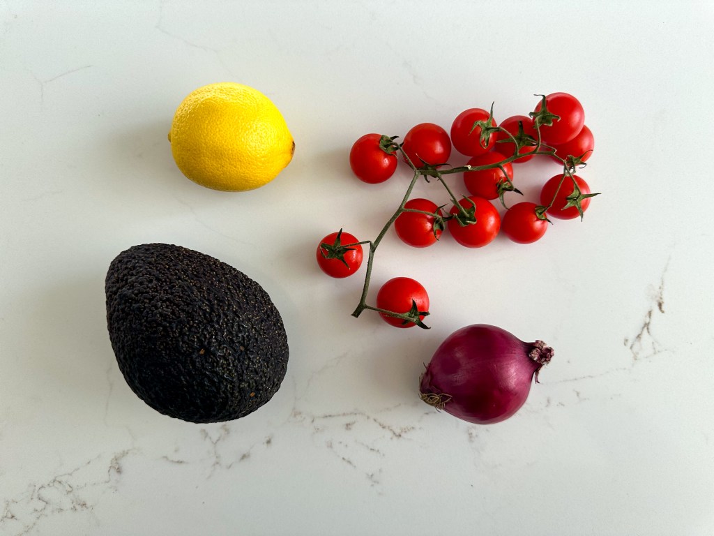 Fresh whole avocado, a lemon, a bunch of cherry tomatoes and a red onion arranged on a white stone surface.