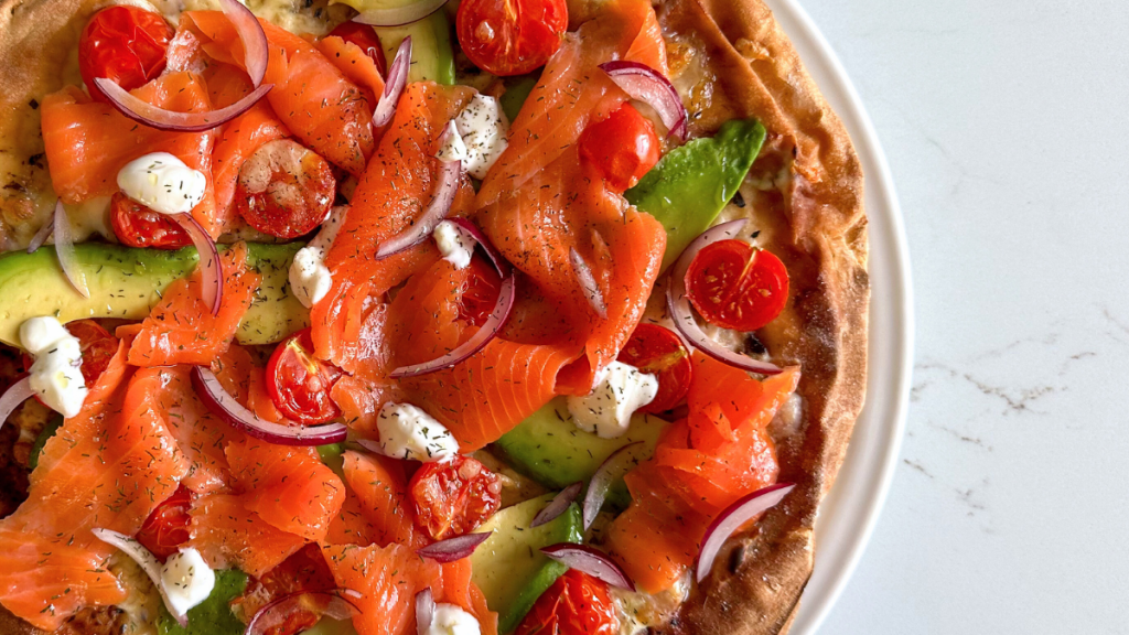 Close-up detail of smoked salmon pizza toppings including red onion slices, cherry tomatoes, avocado and a sprinkle of dill.