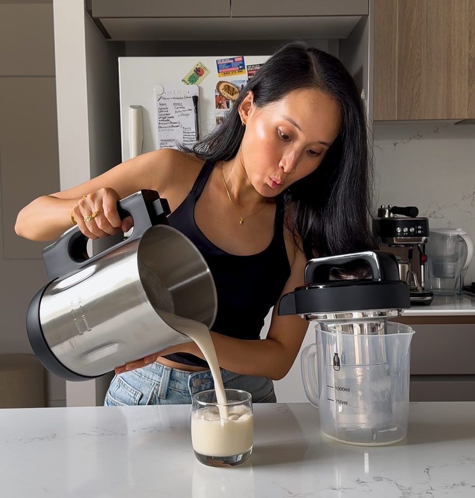 A person pouring creamy, white cashew milk from the Mylky machine jug into a clear glass tumbler on a kitchen bench.