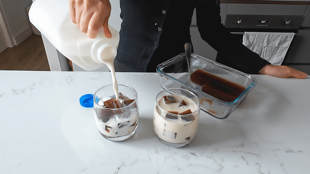 A person pouring white milk from a bottle over dark coffee jelly cubes in a glass.