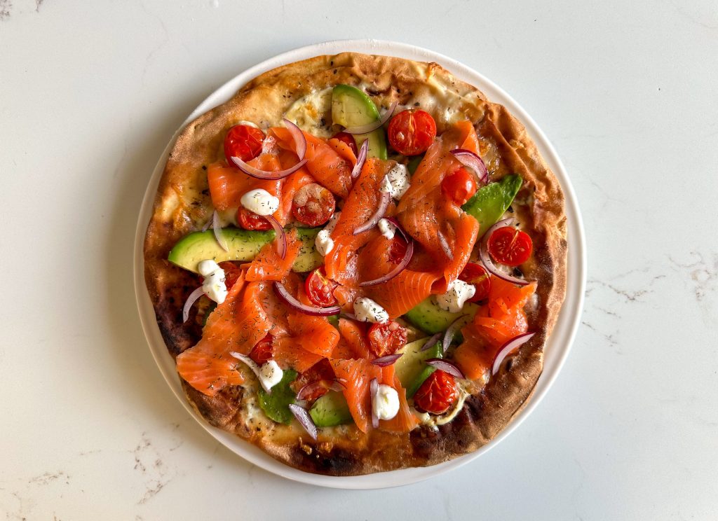 A top-down view of a finished smoked salmon pizza topped with avocado, cherry tomatoes, red onion and Greek yoghurt on a white plate.