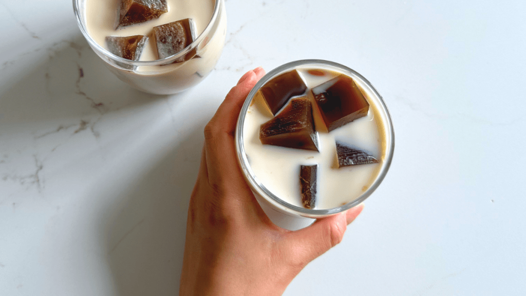 A top-down view of a hand holding a glass filled with dark coffee jelly cubes and creamy milk on a white marble surface.