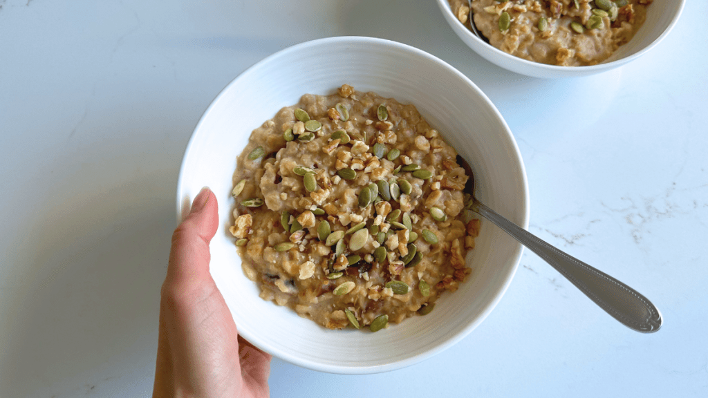 Flat lay view of a hand holding a white bowl of creamy chai-infused oatmeal porridge with chopped dates, topped with green pepitas and crushed walnuts on a white marble surface.