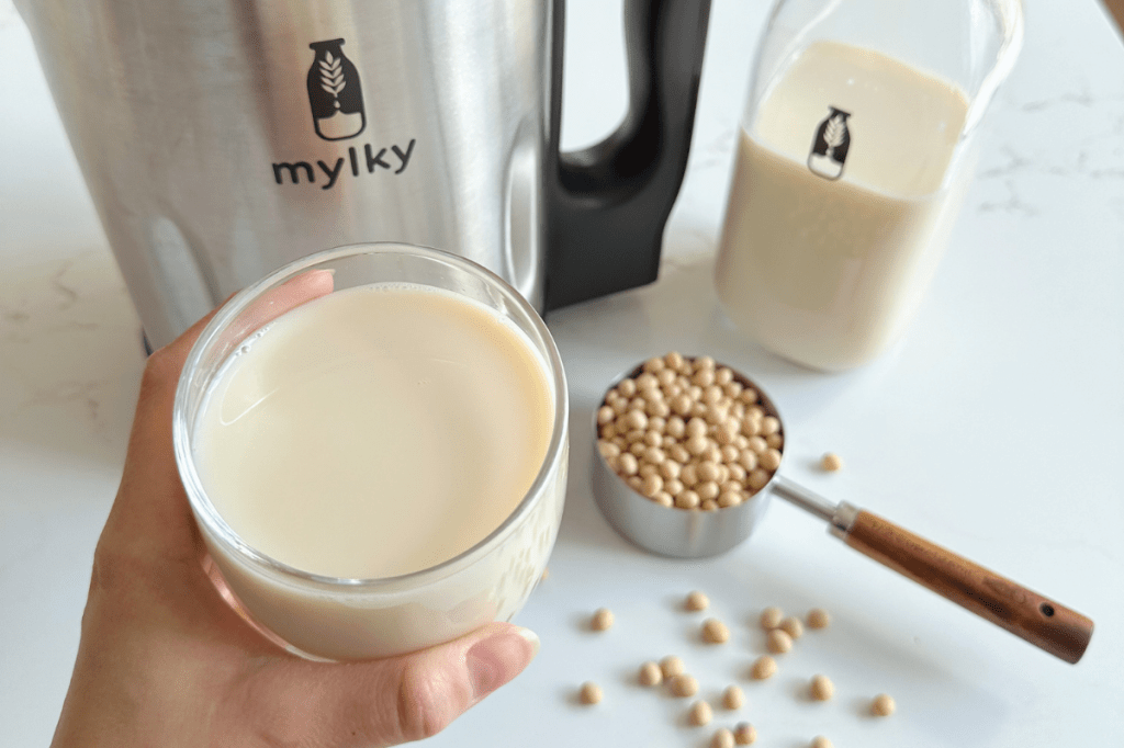 A hand holding a glass of smooth, frothy soy milk in the foreground, with a measuring cup of soybeans and a silver Mylky machine on a white bench top.