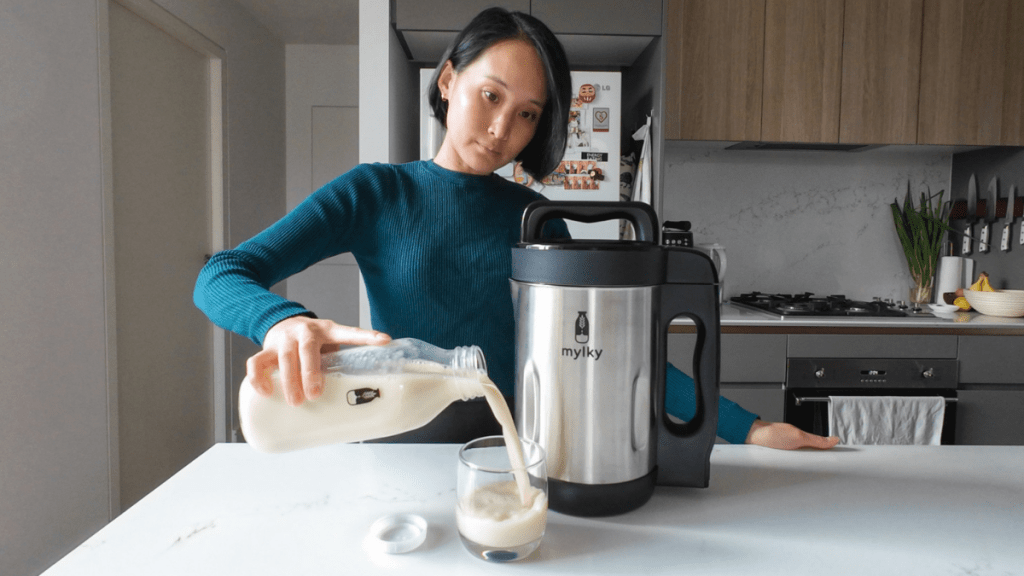 A woman in a teal jumper pouring fresh, creamy white soy milk from a glass bottle into a glass on a white marble kitchen bench next to a silver Mylky machine.