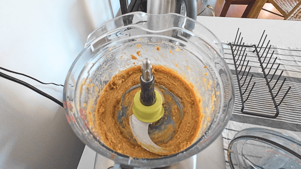 Inside a clear food processor bowl showing a smooth, thick, golden-brown paste made from blended peanut butter, honey, and dates.