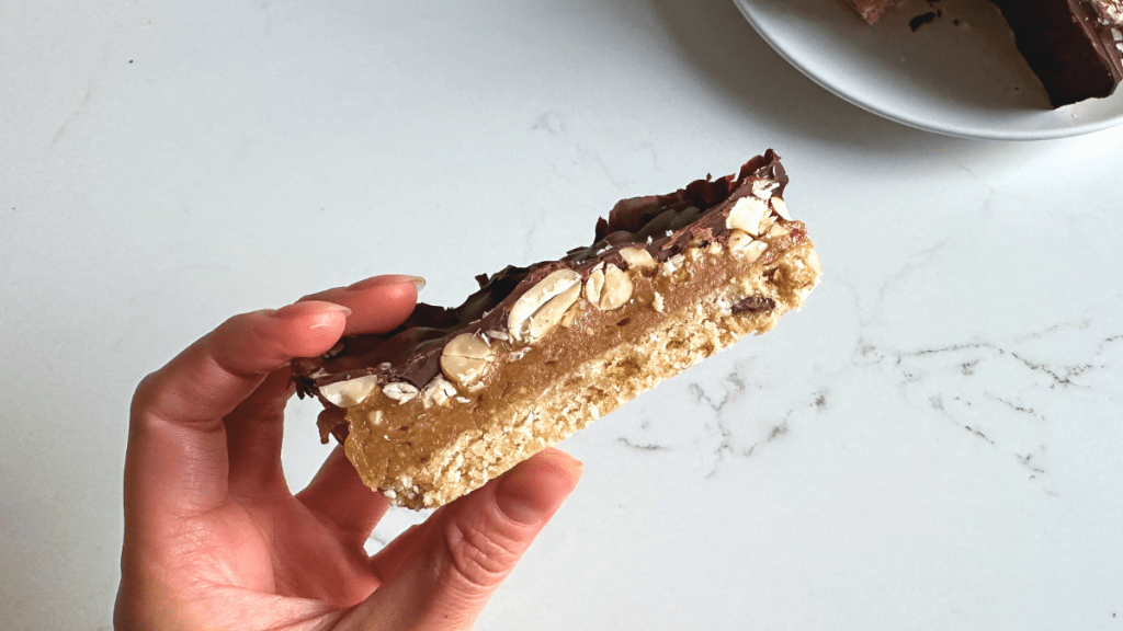 A hand holding a thick, four-layered no-bake chocolate nut bar showing the distinct coconut base, gooey caramel, crunchy whole peanuts, and dark chocolate coating.