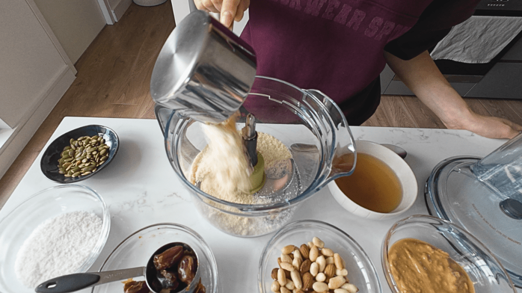 Pouring almond meal from a stainless steel measuring cup into a food processor bowl to make the raw biscuit crust layer.