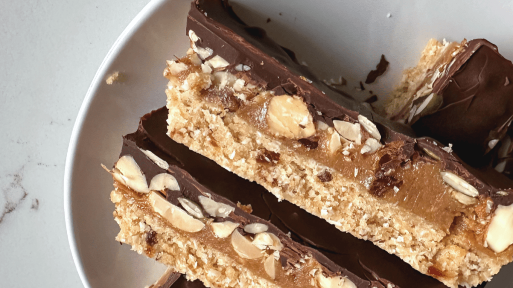 A close-up overhead view of sliced dark chocolate caramel nut bars stacked in a white bowl, showing embedded almonds and pepita seeds.