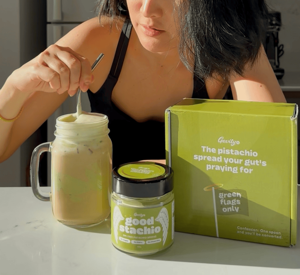 A woman stirring a glass mason jar of iced pistachio latte topped with thick pistachio cold foam using a stainless steel straw, sitting next to a jar and box of Goodstachio Pistachio Spread on a counter.