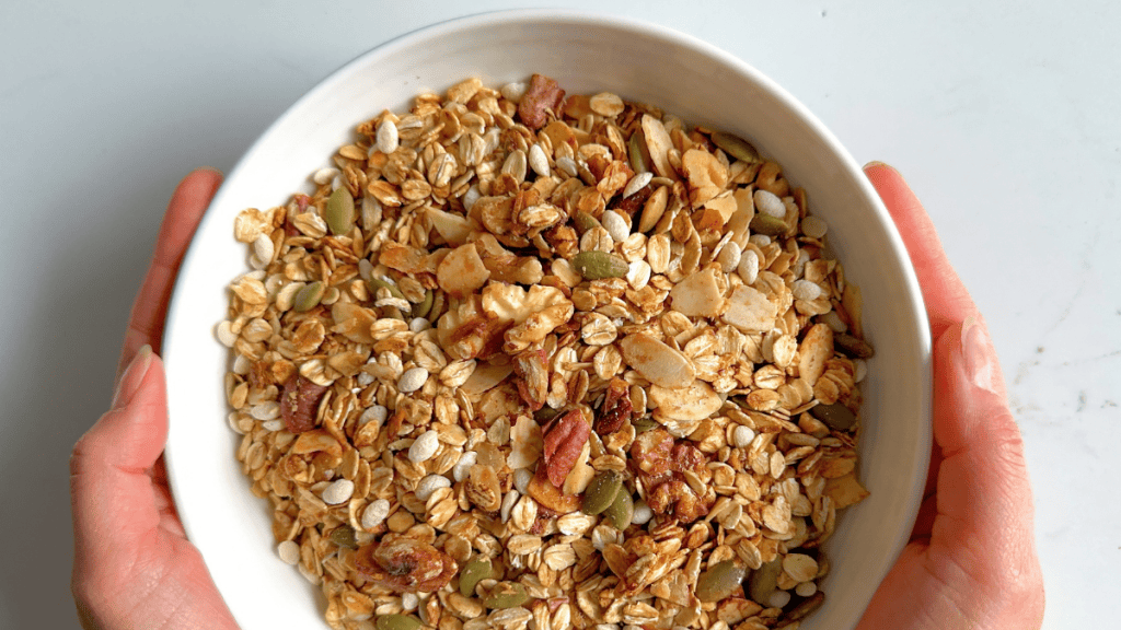 Hands holding a white bowl filled with golden toasted muesli with nuts, seeds, and puffed rice.